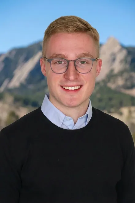 Antoni Czolgowski, smiling, in glasses, a light blue shirt and a black sweater, with mountains behind him